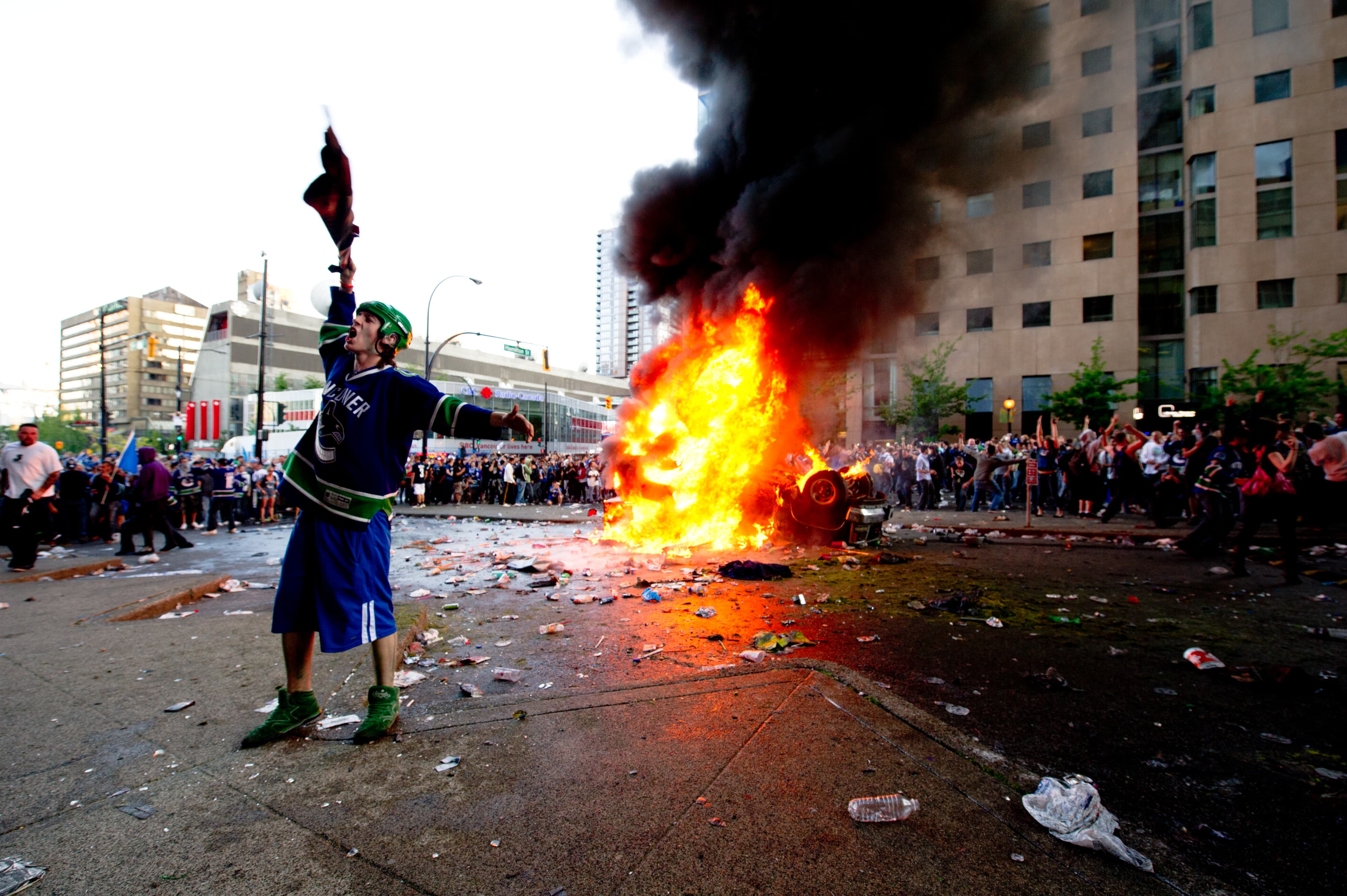 A person in a Vancouver hockey jersey stands near a burning car during unrest in downtown Vancouver.